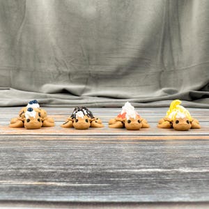 May include: Four miniature turtle figurines with tan bodies and various toppings. One has a blue and white topping, another has black and white, a third has red and white, and the last has yellow and white. The turtles are on a wooden surface.