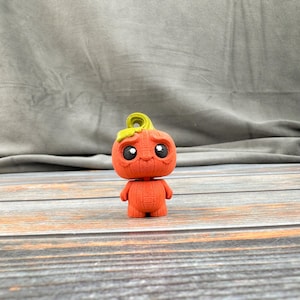 May include: A small, orange pumpkin-shaped figurine with a sad expression. The figurine has large black eyes, a green stem, and small arms. It is standing on a wooden surface against a gray background.
