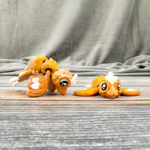 May include: Two whimsical, orange and white bird figurines with large, expressive eyes. One bird is upright, while the other is lying down. The figurines are on a wooden surface, with a gray fabric backdrop.