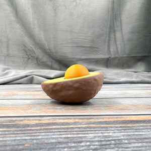 May include: A halved avocado with a yellow yolk-like object inside. The avocado is on a wooden surface.