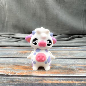 May include: A white cow figurine with purple and pink accents. The toy cow has large black eyes, a pink nose, and purple spots. The ears are pink with purple tips. The belly has a pink circle with a flower design.