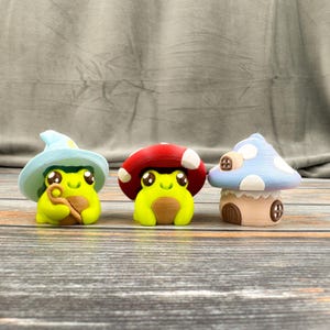May include: Three whimsical frog figurines. One frog wears a wizard hat, another a red mushroom cap, and the third a blue mushroom house. The frogs are yellow and have large, expressive eyes. The background is a neutral gray.