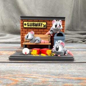 May include: A whimsical diorama featuring three cartoon rats in a subway scene. The scene includes a brick wall with a "SUBWAY" sign, a bench, a trash can, and miniature objects. The rats are gray with pink tails and ears.