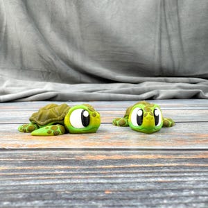 May include: Two small green and brown turtle figurines with large black eyes. The turtles are facing each other on a wooden surface.