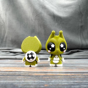 May include: Two green alien-like figurines with large black eyes and star-shaped pupils. One has a round head and a soccer ball design on its chest. The other has antennae and a backpack. Both have white and green outfits.