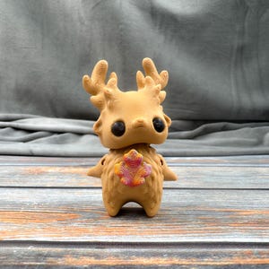 May include: A tan-colored figurine of a creature with antlers, large black eyes, and small arms. A colorful, patterned design is on its chest. The figurine is standing on a wooden surface against a gray background.