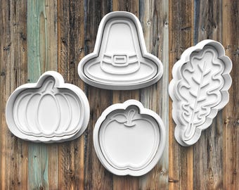 May include: Four white plastic cookie cutters in the shapes of a pumpkin, a pilgrim hat, an apple, and a leaf. The cutters are arranged on a wooden background with vertical planks of varying shades of brown and green.