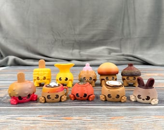May include: A selection of vibrant, quirky food-themed figurines. The set features a popsicle, corn on the cob, a vase, a cupcake, a mushroom, a pumpkin, a cinnamon roll, and a bunny, each with charming faces. They are displayed on a wooden surface.