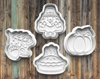 May include: Four white plastic cookie cutters in Thanksgiving shapes: a turkey wearing a pilgrim hat, a pumpkin, a pie, and an acorn with oak leaves. The cutters are on a wooden background.