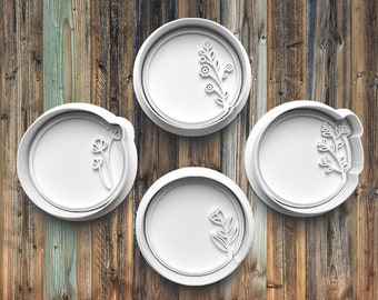 May include: Four white, circular cookie cutters with floral designs. Each cutter has a different embossed botanical pattern, including sprigs and flowers. The cutters are arranged on a rustic, wooden background.