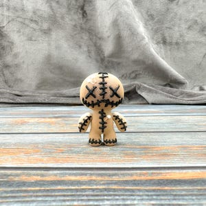 May include: A small, beige voodoo doll with black stitching around the eyes and mouth. The doll has a stitched-on smile and X's for eyes.