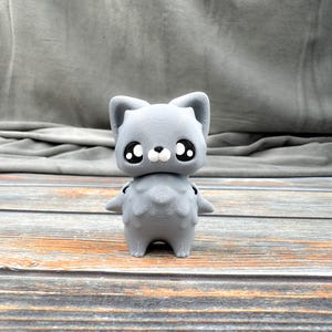 May include: A gray 3D-printed cat figurine with large black eyes and small arms. The cat has a textured surface and is standing on a wooden surface. The figurine has a cute, cartoonish appearance.