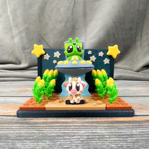 May include: A whimsical diorama featuring a green alien in a UFO above a cow. The scene includes corn stalks, stars, and a blue backdrop. The base is brown and the UFO is gray and blue.