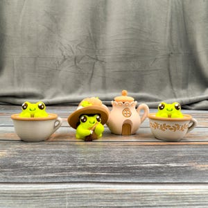 May include: A whimsical collection of frog figurines and a teapot. Two frogs sit in teacups, one wears a hat and holds a broom, and the teapot has a door. The figurines are green, beige, and brown, creating a charming, fairytale aesthetic.