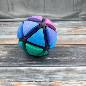 May include: A spherical geometric puzzle toy featuring a dark purple frame with multiple triangular inserts in shades of light purple, pink, blue, and teal. Each triangular piece displays a concentric circular pattern on its surface.