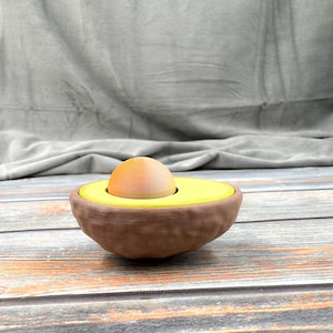 May include: A brown avocado-shaped egg holder with a yellow avocado pit and a brown egg inside.