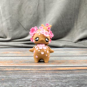 May include: A brown figurine with a floral design. The figurine has a round body, small arms, and large eyes. Pink flowers adorn the head and body. The figurine is standing on a wooden surface.