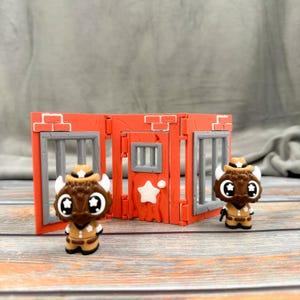 May include: Two miniature bison figures wearing cowboy hats and outfits stand in front of a red jail set with gray bars and a door with a star. The bison figures have large eyes and are holding small guns.