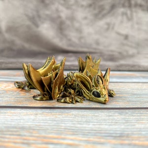 May include: A gold-coloured 3D-printed dragon figurine. The dragon has large ears, a detailed face, and textured wings. The figurine is resting on a wooden surface.