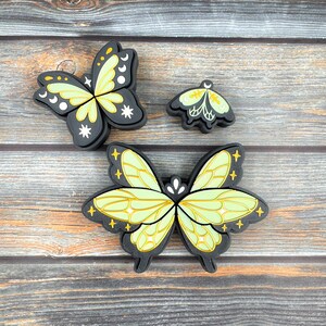 May include: Three butterfly-shaped decorative items. The largest butterfly has light green wings with black outlines and yellow accents. The other two butterflies are smaller, with similar designs. The butterflies are on a wooden surface.