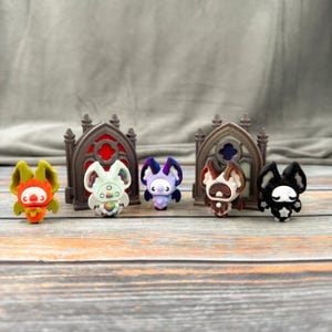 May include: Five small bat figurines in various colors including orange, white, purple, brown, and black, each with distinct facial markings. Two dark gray gothic-style archway structures with red and blue inserts stand behind the bat figures.