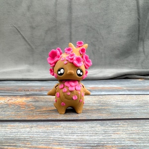 May include: A brown figurine of a creature with large eyes and pink flower-like accents on its head and body. The figurine has small arms and a rounded body, with a cute and whimsical design. The background is a gray fabric and a wooden surface.