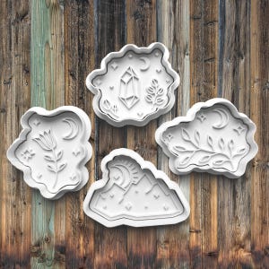 May include: Four white cookie cutters with different designs: a crystal, a mountain range, a flower, and a branch. The cutters are on a wooden background.