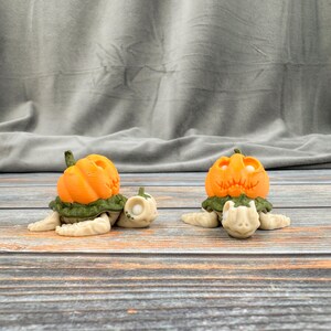 May include: Two decorative turtle figurines with orange pumpkin heads and green leafy collars. The turtles are cream-coloured with detailed features, and one has a small pumpkin beside it. The figurines are on a wooden surface.
