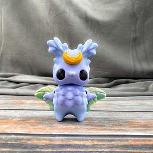 May include: A light blue figurine with a yellow crescent moon on its head and large black eyes. The figurine has detailed wings with green and yellow patterns. The figure is standing on a wooden surface.