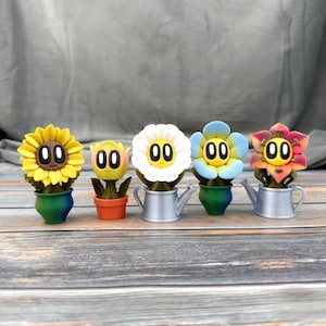 May include: Five whimsical flower figurines with cartoon faces. The flowers are yellow, white, blue, and red-orange, each in a small pot or watering can. The pots are green, orange, and silver. The background is a gray fabric and a wooden surface.