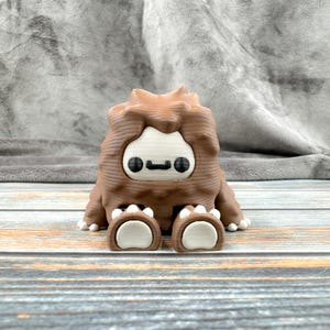 May include: A brown and white 3D printed figurine of a creature with a furry brown body, white face, and black eyes. The creature is sitting with its legs crossed and has white paws.