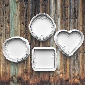 May include: Four white plastic cookie cutters in various shapes: a circle with a scalloped edge, a round frame with heart accents, a square frame with heart accents, and a heart shape. The cutters are arranged on a wooden background.