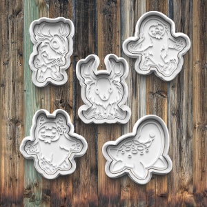 May include: Five white cookie cutters shaped like different animals. The cutters are on a wooden background. The animals are a deer, a sloth, a turtle, a monster with flowers, and a monster with horns.