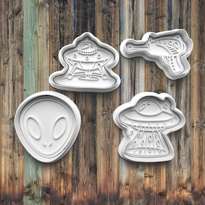 May include: Four white plastic cookie cutters in an alien theme. The shapes include a classic alien head, a ray gun, and two different flying saucer designs. The cutters are set against a wooden background.