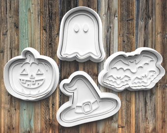 May include: Four white Halloween-themed cookie cutters. The shapes include a jack-o'-lantern, a ghost, a witch's hat, and a cluster of bats. The cutters are made of plastic and are set against a wooden background.