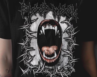 May include: A black short-sleeve t-shirt featuring a graphic print of an open mouth with sharp fangs, surrounded by a rectangular frame of barbed wire. The text Blood Moon is printed in a stylized, jagged black font above the graphic.