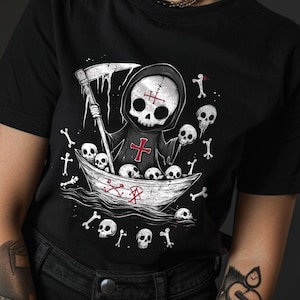 May include: Black t-shirt with a graphic of a small skeleton wearing a hooded robe and holding a scythe. The skeleton is in a boat with a skull and crossbones design. The boat is surrounded by bones and skulls.