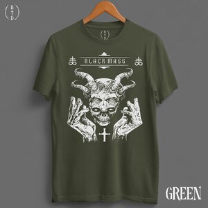 May include: An olive green short-sleeve t-shirt featuring a white graphic print of a horned skull with hands raised on either side. The text "BLACK MASS" is printed above the skull, flanked by two cross symbols. A small cross is below the skull.