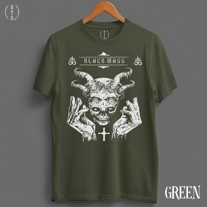 May include: An olive green short-sleeve t-shirt featuring a white graphic print of a horned skull with hands raised on either side. The text "BLACK MASS" is printed above the skull, flanked by two cross symbols. A small cross is below the skull.