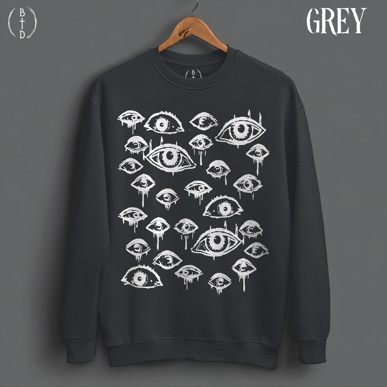 May include: A dark grey crewneck sweatshirt featuring a white graphic print of numerous eyes of varying sizes. Many of the eyes have dripping lines extending downward. The word GREY is printed in the upper right corner, and a logo is in the upper left.