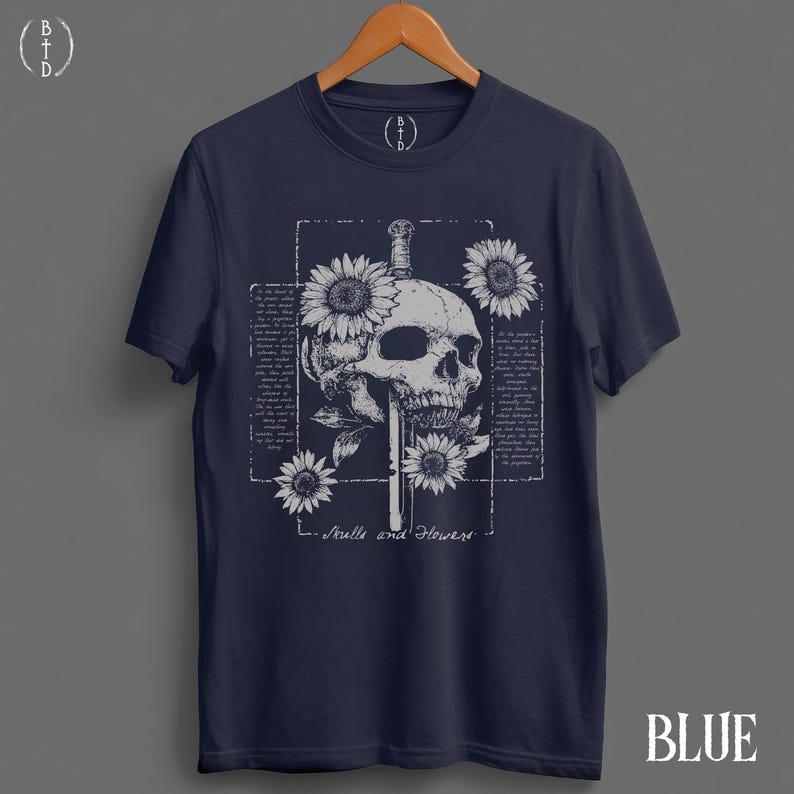 May include: Navy blue t-shirt featuring a detailed skull design pierced by a sword, surrounded by sunflowers. The graphic includes the text "Skulls and Flowers" and is set within a rectangular frame. The word "BLUE" is at the bottom.