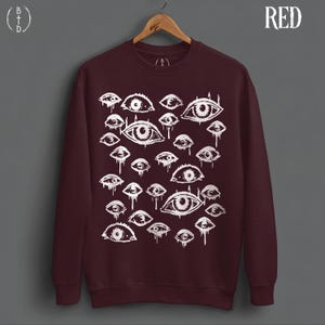 May include: A dark maroon crewneck sweatshirt featuring a white graphic print of numerous eyes of varying sizes. The eyes appear to be dripping or weeping. The sweatshirt is displayed on a wooden hanger against a gray background. The word RED is printed in white in the top right corner.