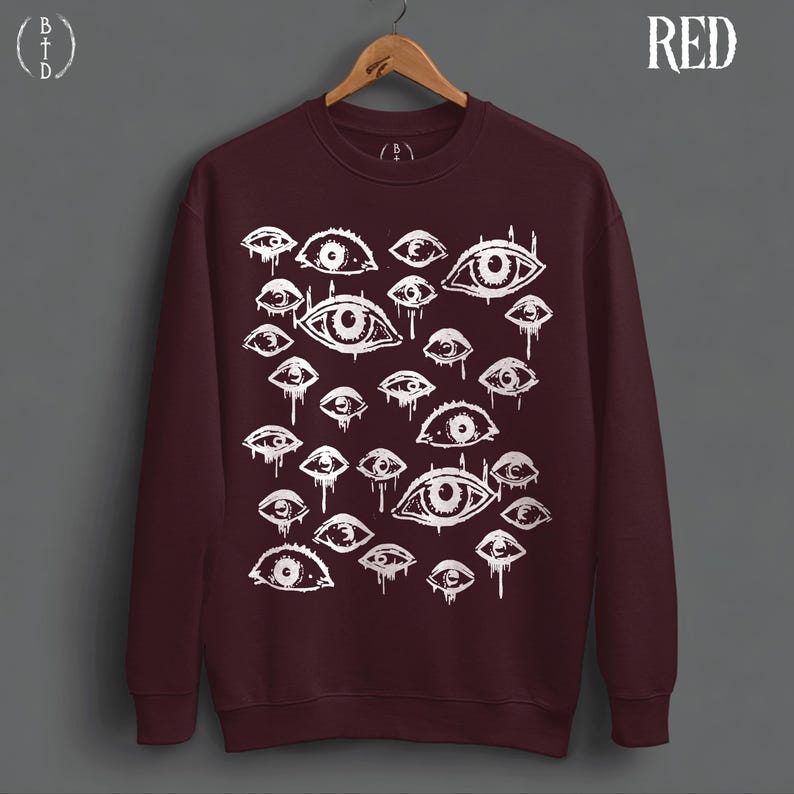 May include: A dark maroon crewneck sweatshirt featuring a white graphic print of numerous eyes of varying sizes. The eyes appear to be dripping or weeping. The sweatshirt is displayed on a wooden hanger against a gray background. The word RED is printed in white in the top right corner.