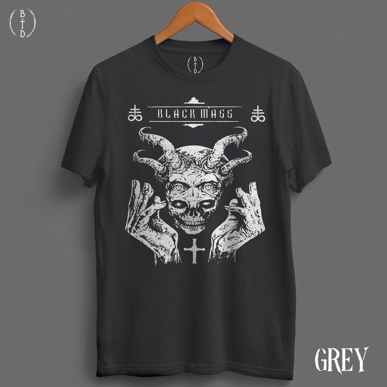 May include: A charcoal grey short-sleeve t-shirt featuring a white graphic print of a horned skull flanked by two hands. The text "BLACK MASS" is printed above the skull, accompanied by two cross symbols and a small cross below the chin.