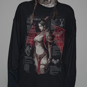 May include: A black long-sleeve shirt featuring a graphic print of a succubus with red wings. The design includes technical-style text, a heart illustration, and various symbols surrounding the central figure, all rendered in shades of red and grey.