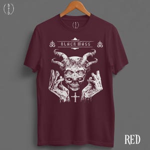 May include: A maroon short-sleeve t-shirt featuring a white graphic print of a horned skull with hands raised on either side. The text "BLACK MASS" is printed above the skull, flanked by two cross symbols. A small cross is centered below.