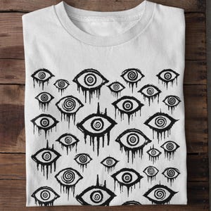 May include: White t-shirt featuring a black and white print of numerous stylised eyes with swirling patterns. The eyes are dripping with black paint.