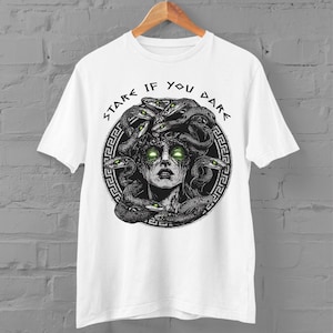 Op de afbeelding: Wit T-shirt met een zwart-witte illustratie van Medusa met slangen als haar. De tekst "Stare If You Dare" is geschreven in een cirkelvormig ontwerp rond Medusa's hoofd.