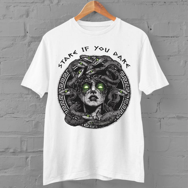 Op de afbeelding: Wit T-shirt met een zwart-witte illustratie van Medusa met slangen als haar. De tekst "Stare If You Dare" is geschreven in een cirkelvormig ontwerp rond Medusa's hoofd.