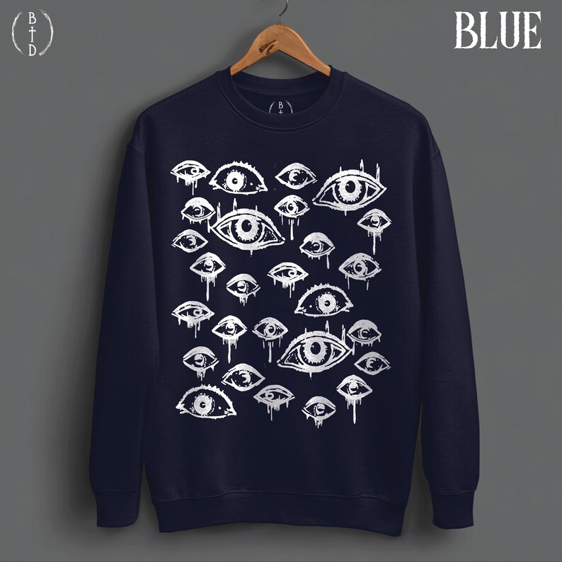 May include: A navy blue crewneck sweatshirt featuring a white graphic print of numerous eyes of varying sizes. Many of the eyes have a dripping effect. The word BLUE is printed in the top right corner, and a BTD logo is in the top left.